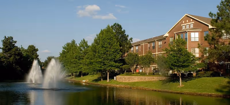 Brick senior living building with green lawns, trees, and a pond with fountains