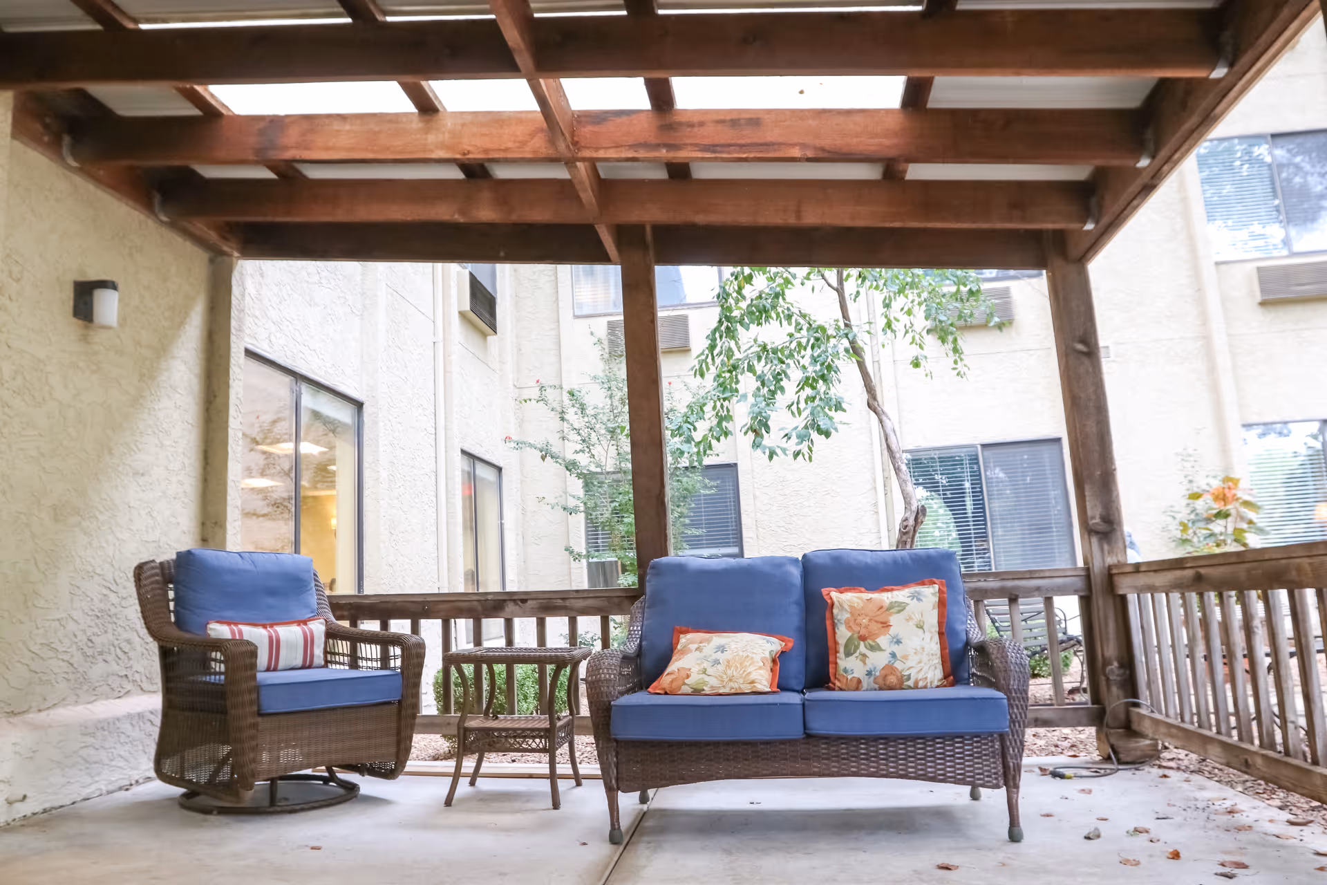Covered outdoor seating area with wicker sofa and chairs with blue cushions under a wooden pergola in a courtyard.
