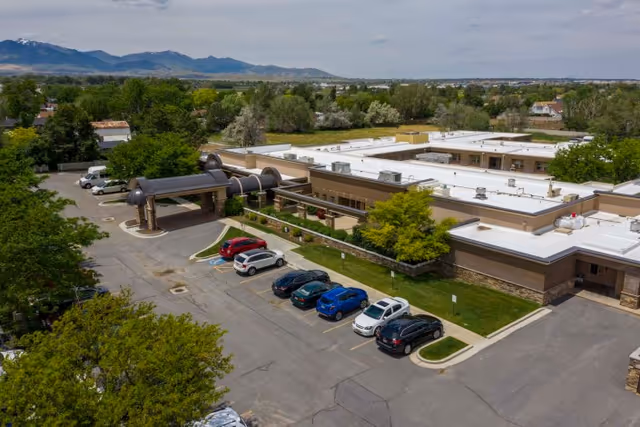 Aerial view of Copper Ridge Health Care facility showing a single-story building with a covered entrance, several parked cars in front, surrounded by trees and greenery with mountains visible in the background under a partly cloudy sky.
