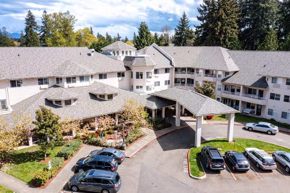 Aerial view of Solstice Senior Living at Renton building showing a large, multi-story facility with a covered entrance driveway, surrounded by trees and parked cars in the parking lot under a partly cloudy sky.