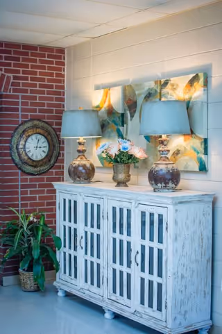 A cozy corner with a distressed white wooden cabinet topped with two rustic table lamps and a vase of flowers. Behind the cabinet is a colorful abstract painting on a white brick wall, and to the left is a round wall clock mounted on a red brick wall. A green potted plant in a woven basket sits on the floor next to the cabinet.