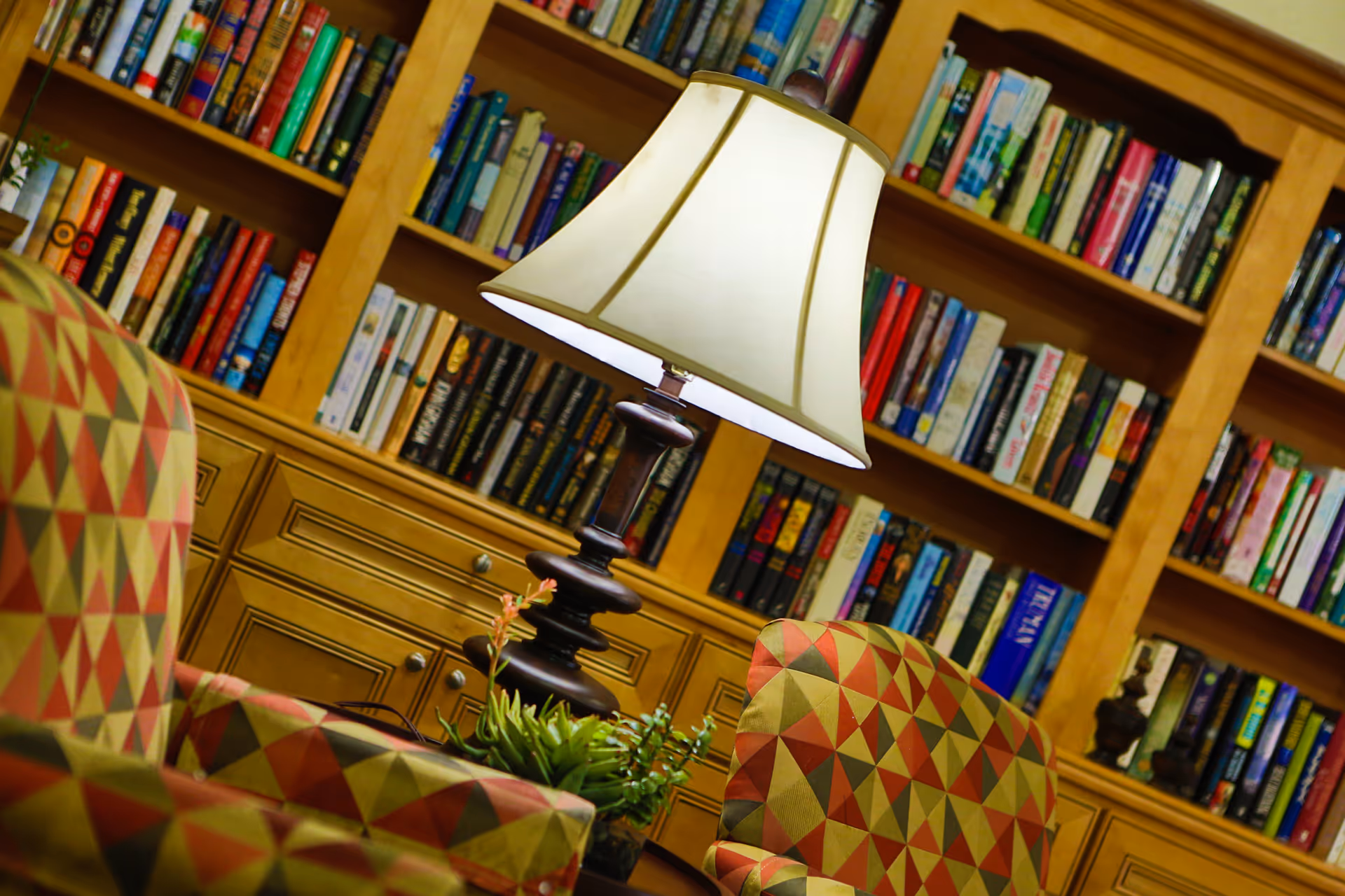 A cozy reading area with two patterned armchairs, a table with a lamp, and a wooden bookshelf filled with books in the background.
