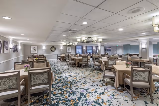 A spacious dining room with multiple tables covered in beige tablecloths, each surrounded by cushioned chairs. The room features a patterned carpet with blue, green, and orange accents, soft overhead lighting, and framed artwork on the walls. Small flower arrangements are placed on the tables.