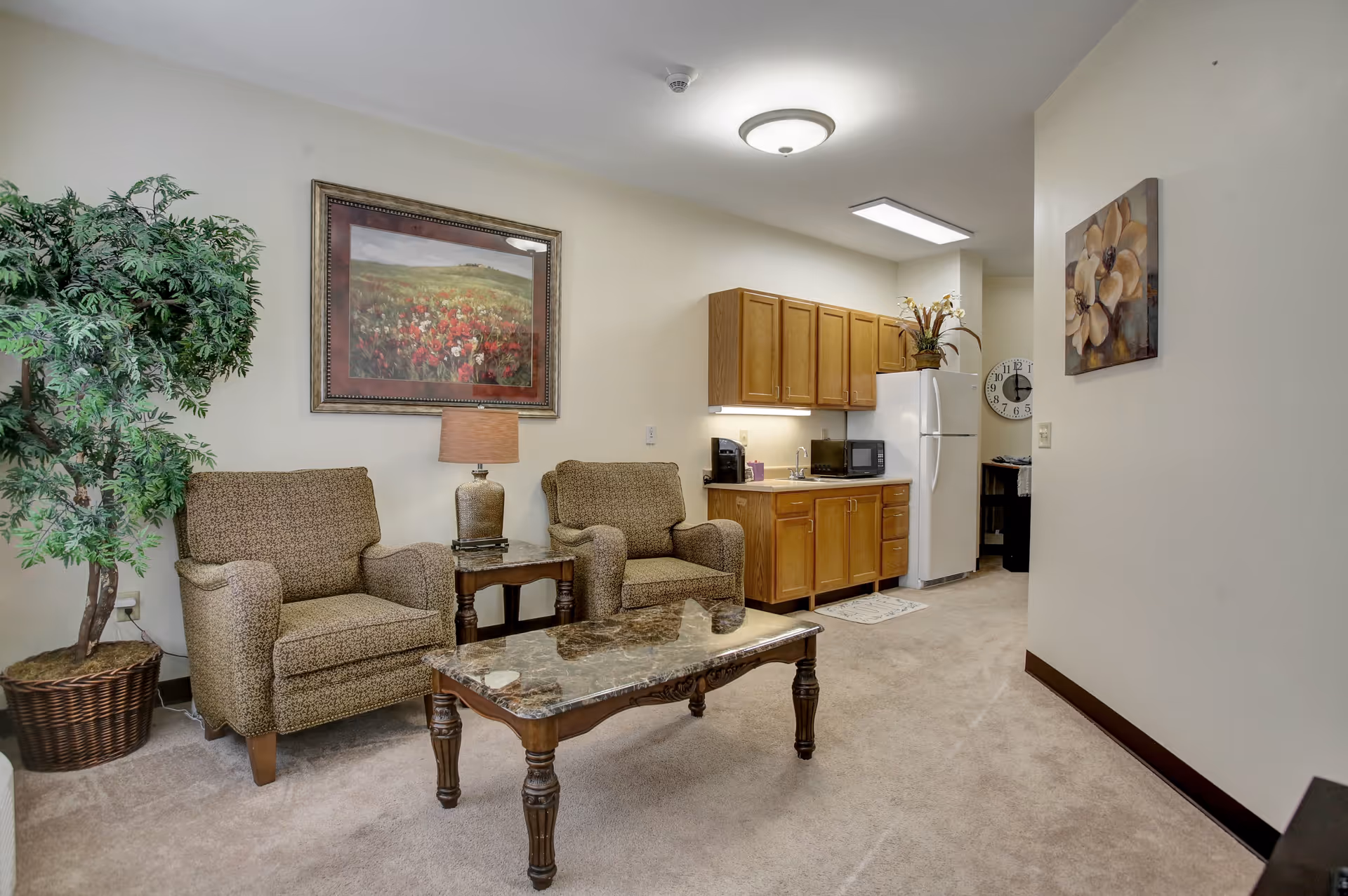 A cozy living area with two patterned armchairs separated by a small wooden side table with a lamp. A large framed painting of a flower field hangs on the wall above the chairs. To the right, there is a small kitchenette with wooden cabinets, a white refrigerator, a microwave, and a coffee maker. A decorative plant is placed on top of the refrigerator, and a large potted plant is positioned on the left side of the chairs. The room has beige carpet and neutral-colored walls with additional floral artwork on the right wall.
