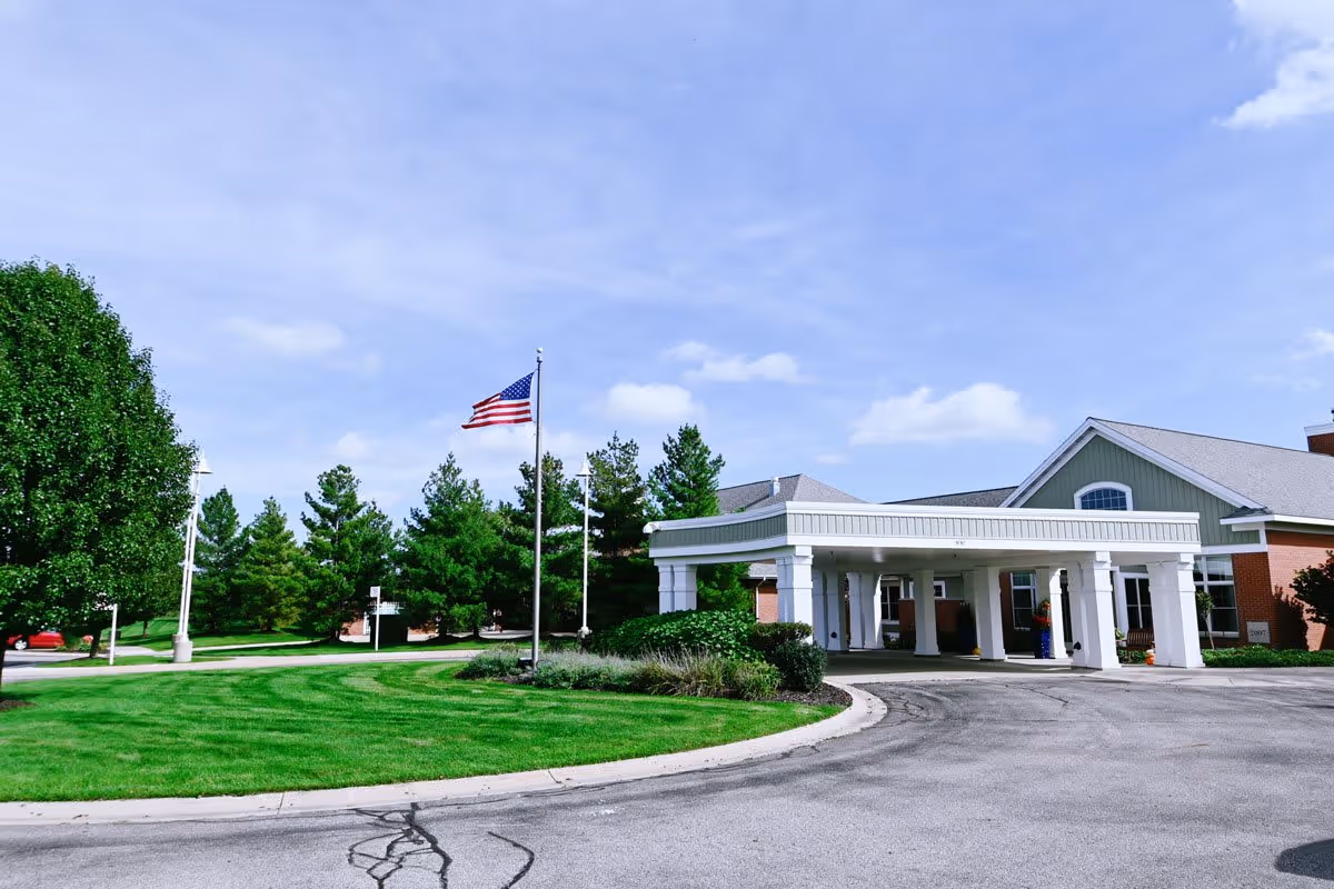 Exterior view of The Cortland Holland Meadows facility showing a driveway with a covered entrance supported by white columns, a well-maintained green lawn, trees, and an American flag on a flagpole under a partly cloudy sky.