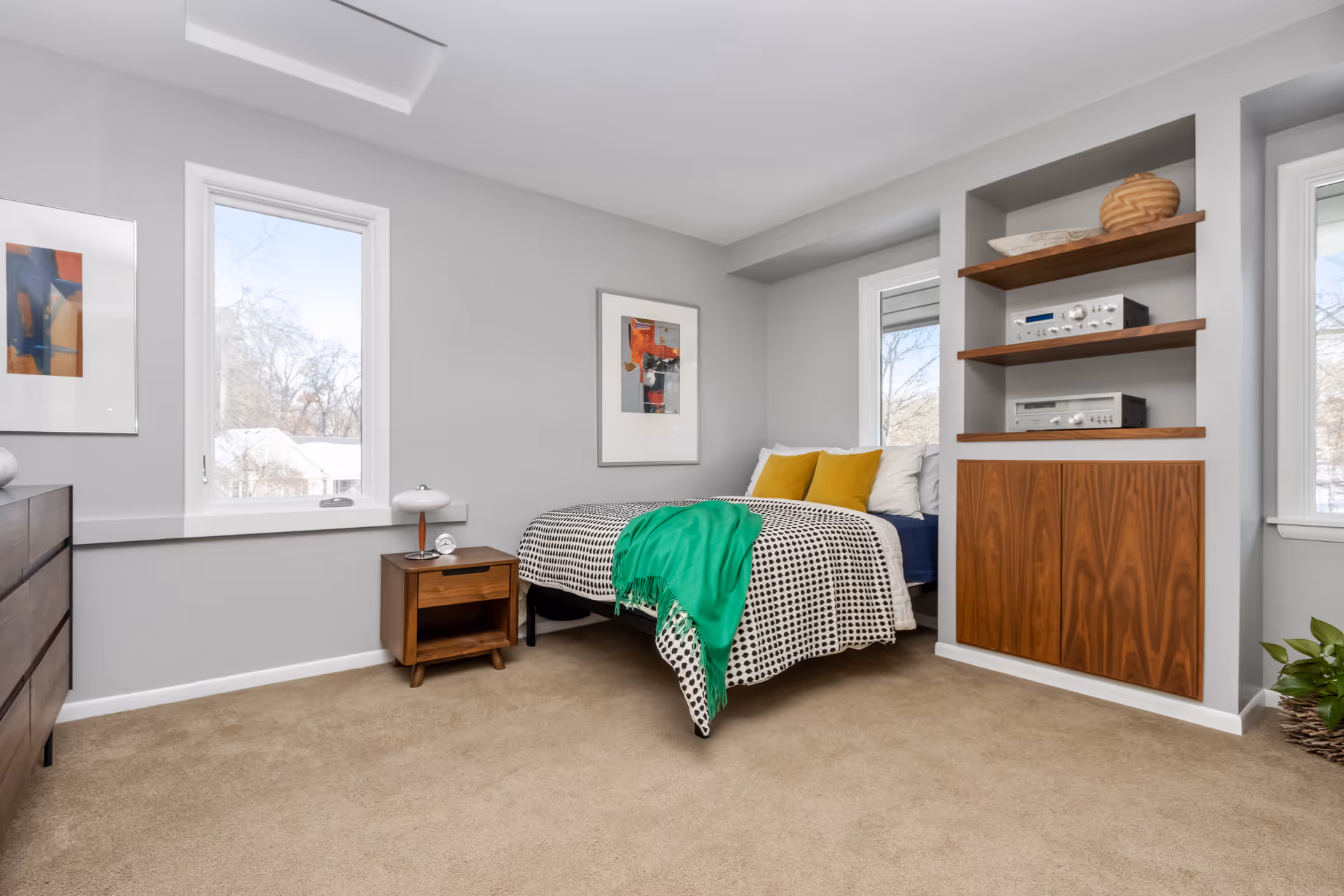A bright bedroom with beige carpet, light gray walls, and two windows showing an outdoor view. The room features a bed with a black and white patterned blanket, yellow pillows, and a green throw. There is a wooden nightstand with a small lamp and clock, a wooden dresser, framed abstract artwork on the walls, and built-in wooden shelves with audio equipment and decorative items.