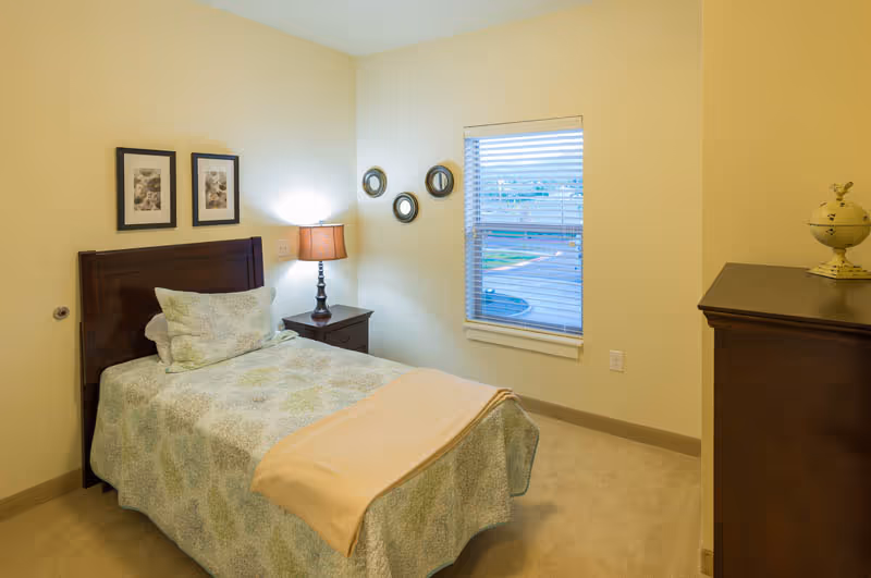 Small furnished bedroom with a single bed, nightstand and lamp, dresser, wall art, and a window.