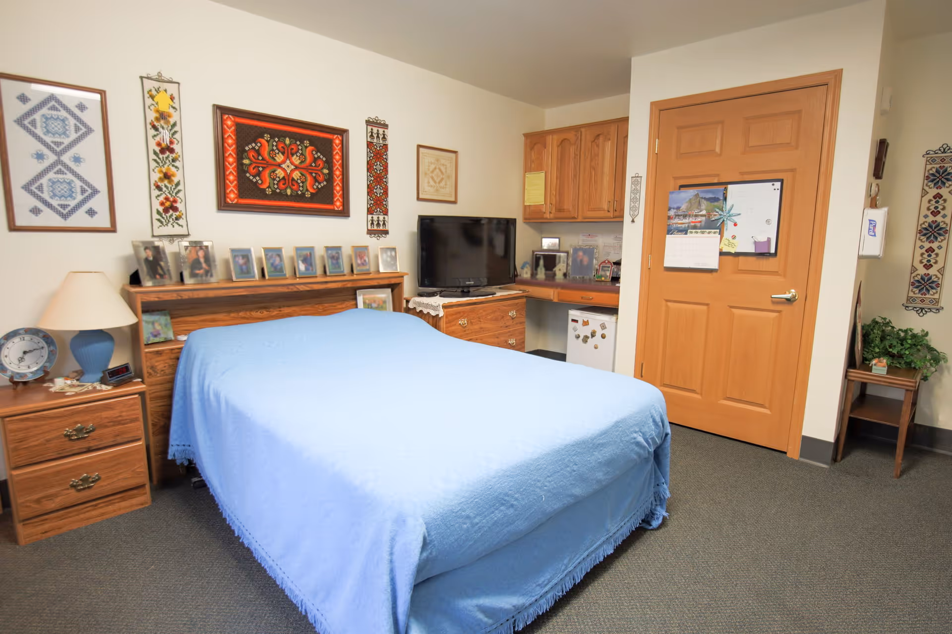 A cozy bedroom in an assisted living facility featuring a bed with a light blue bedspread, a wooden headboard with framed photos, a nightstand with a lamp and clock, a TV on a wooden dresser, wall decorations including framed artwork and embroidered hangings, a wooden door with a calendar and whiteboard, and a small kitchenette area with cabinets and a mini fridge.