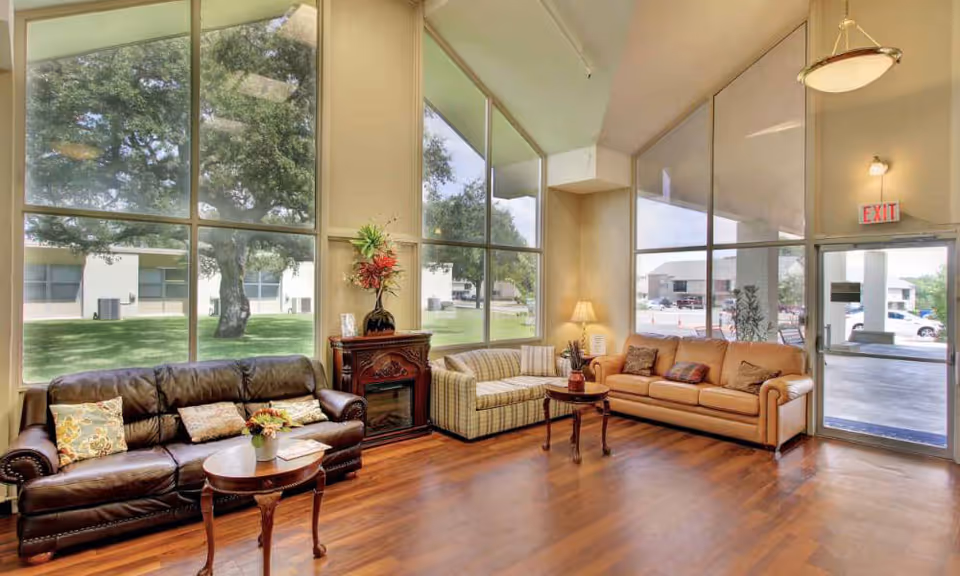 A bright and spacious living room area with large floor-to-ceiling windows showing a green outdoor space with trees. The room features three sofas: one dark brown leather, one beige striped, and one light brown leather. There are two small wooden coffee tables with decorative items, a small lamp on a side table, and a decorative fireplace with a flower arrangement on top. The room has wooden flooring and a glass door leading outside.