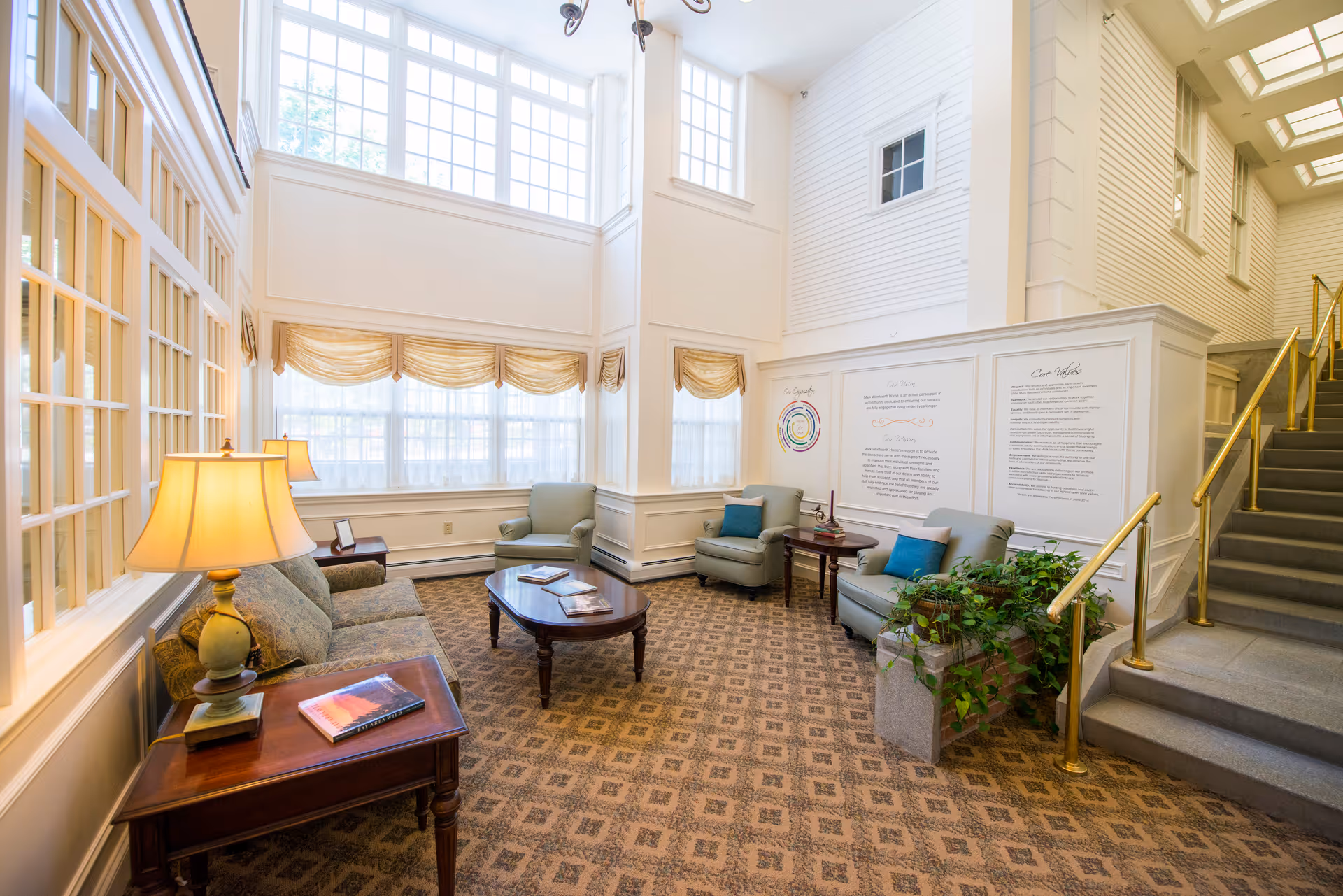 A bright and spacious senior living facility common area with large windows letting in natural light. The room features comfortable armchairs and a sofa arranged around a wooden coffee table. There are side tables with lamps and books, a patterned carpet, and a staircase with brass handrails leading upstairs. The walls are white with decorative paneling and display informational posters.