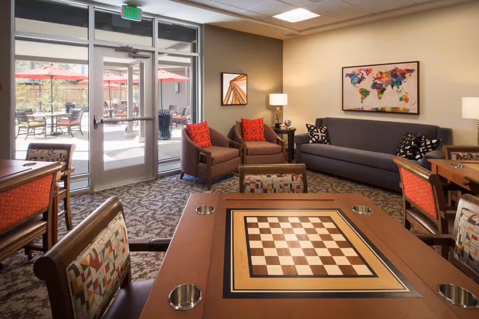 A cozy common lounge with a chessboard game table in the foreground, upholstered chairs and a sofa, and glass doors leading to an outdoor patio with red umbrellas.
