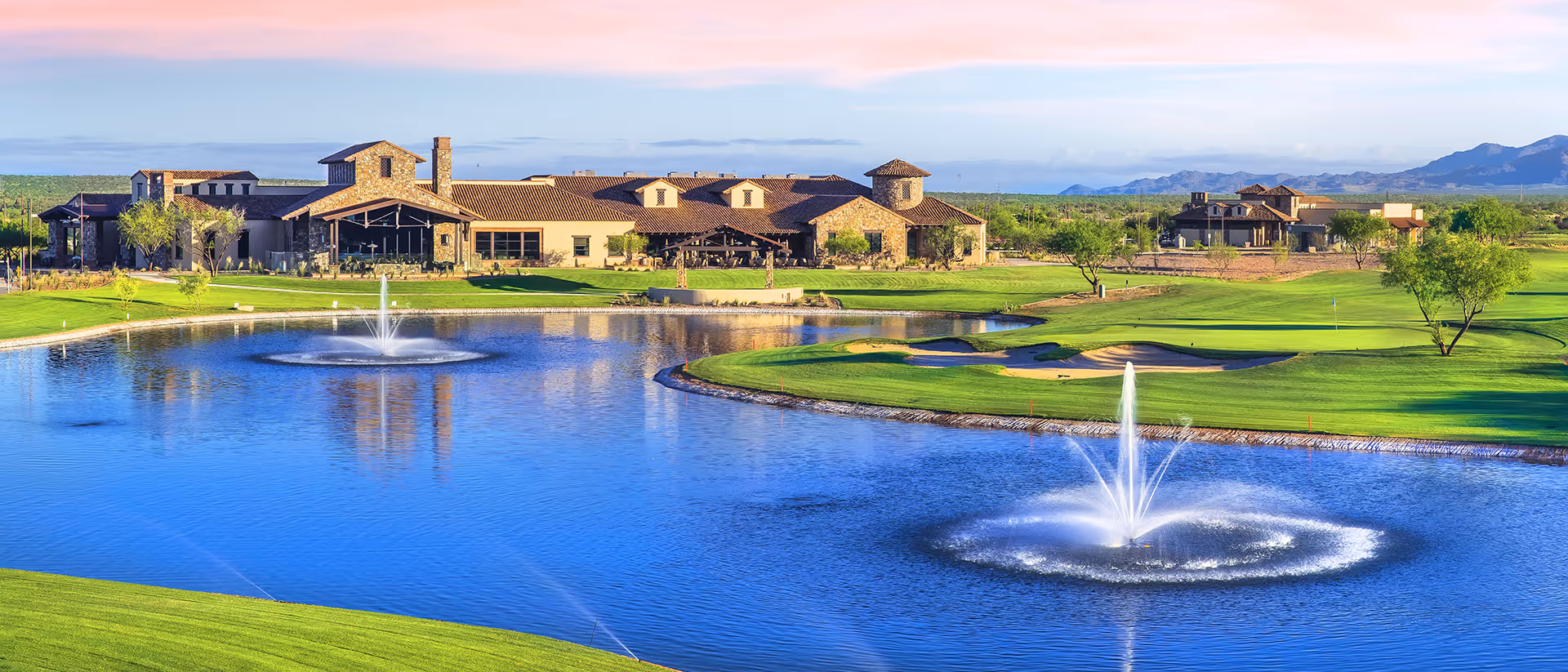 A scenic view of a senior living facility named SaddleBrooke Ranch with a large pond featuring two water fountains in the foreground, surrounded by well-maintained green lawns and golf course areas. The facility buildings are visible in the background with mountains and a clear sky at sunset.