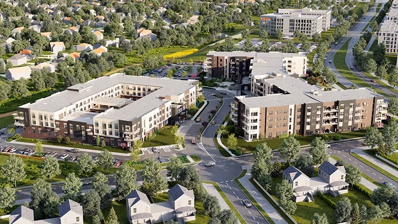 Aerial view of Ovation Heartwood Preserve senior living facility showing multiple modern three-story buildings surrounded by trees, roads, parking lots, and nearby residential houses.