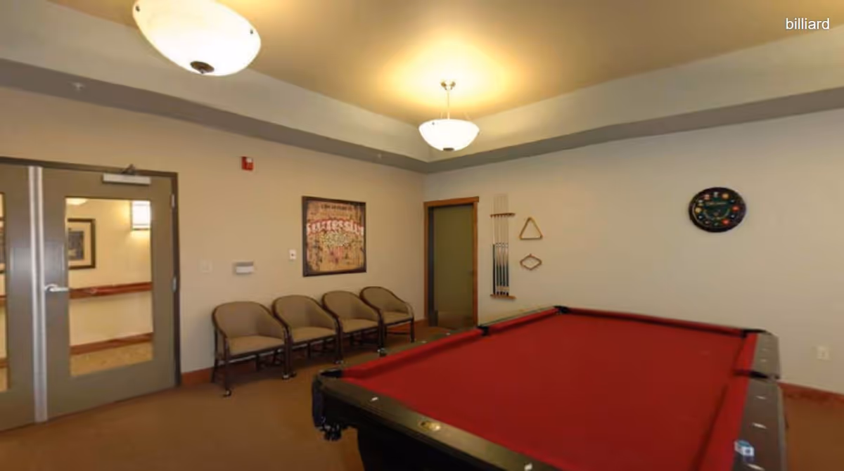 A billiard room with a red pool table in the foreground, four beige chairs lined up against a beige wall, a framed poster, a rack holding pool cues, and a wall-mounted clock shaped like a billiard ball rack. The room has beige walls, a carpeted floor, and two ceiling light fixtures.