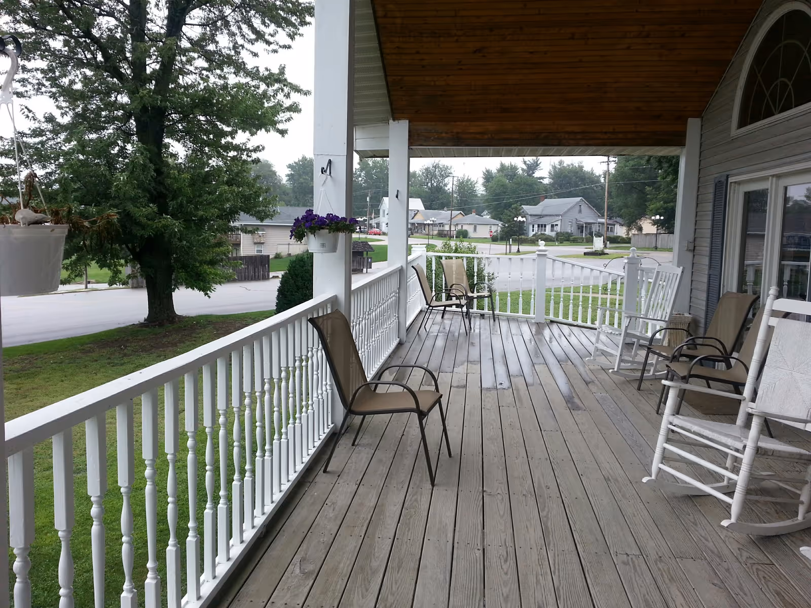 A covered wooden porch with white railings and several chairs including rocking chairs and metal chairs. Hanging flower pots with purple flowers are attached to the porch columns. The porch overlooks a quiet neighborhood street with trees and houses in the background.