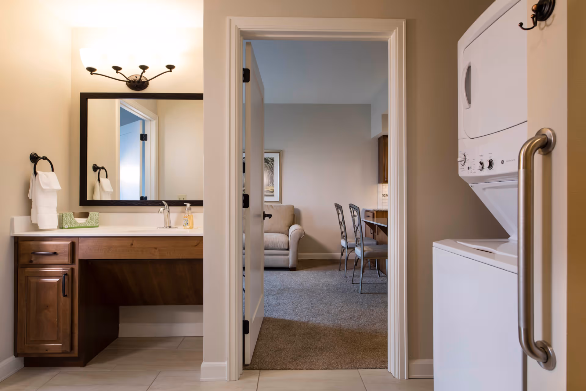 View from a bathroom showing a vanity and mirror on the left, a stacked washer-dryer with grab bar on the right, and a doorway leading to a living area with a sofa and chairs.