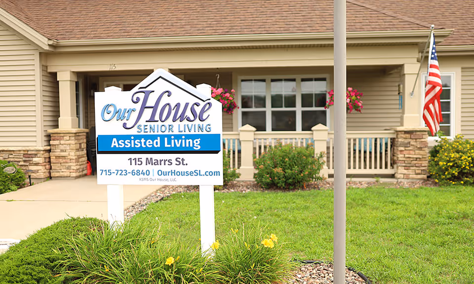 A yard sign reading 'Our House Senior Living Assisted Living' stands in front of a beige senior living home's entrance with landscaping and an American flag.