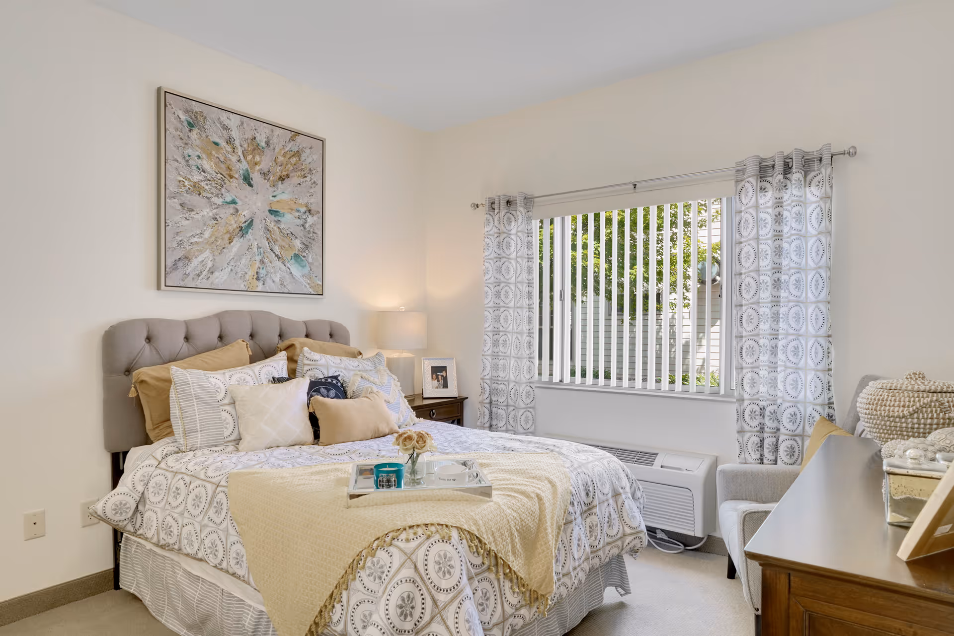 A cozy bedroom with a neatly made bed featuring multiple pillows and a patterned bedspread. There is a bedside table with a lamp and a framed photo, a window with vertical blinds and patterned curtains, and a cushioned chair next to a wooden dresser with decorative items on top. A piece of abstract art hangs above the bed.