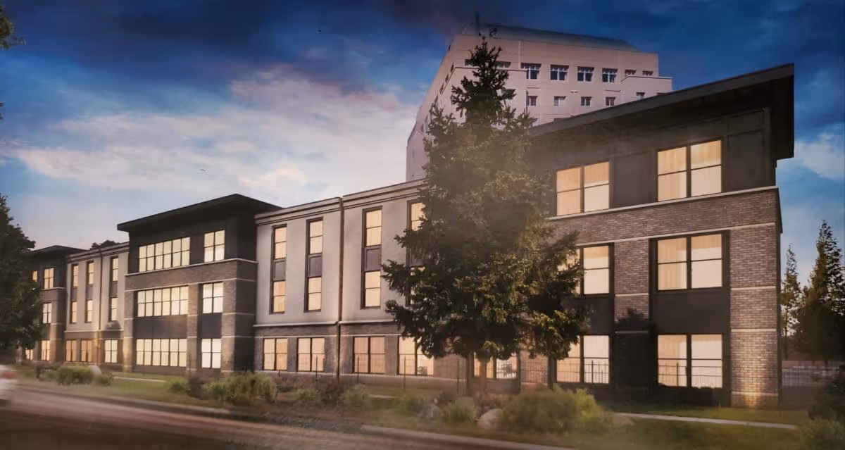 Front exterior of a three-story brick-and-stucco senior living building at dusk with illuminated windows and landscaping.