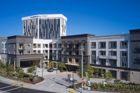 Front exterior of the Cogir of South Bay building showing the main entrance, landscaped driveway, and a tall 'California Bank & Trust' tower in the background.