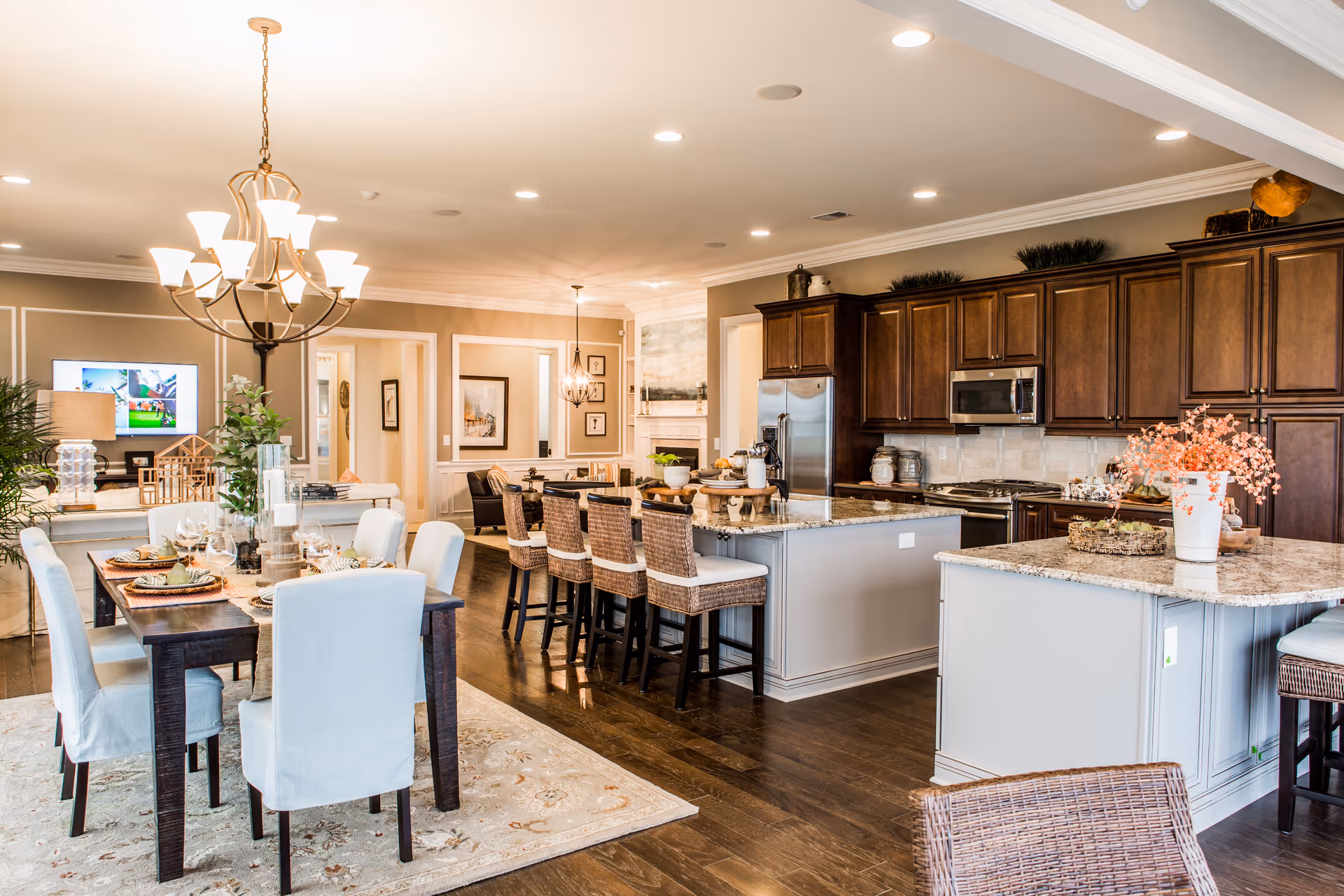 Bright and spacious open-concept kitchen and dining area with dark wood floors, a large island with granite countertops and wicker bar stools, dark wood cabinets, stainless steel appliances, and a dining table set with white upholstered chairs and tableware under a chandelier. The background shows a living area with a TV and additional seating.