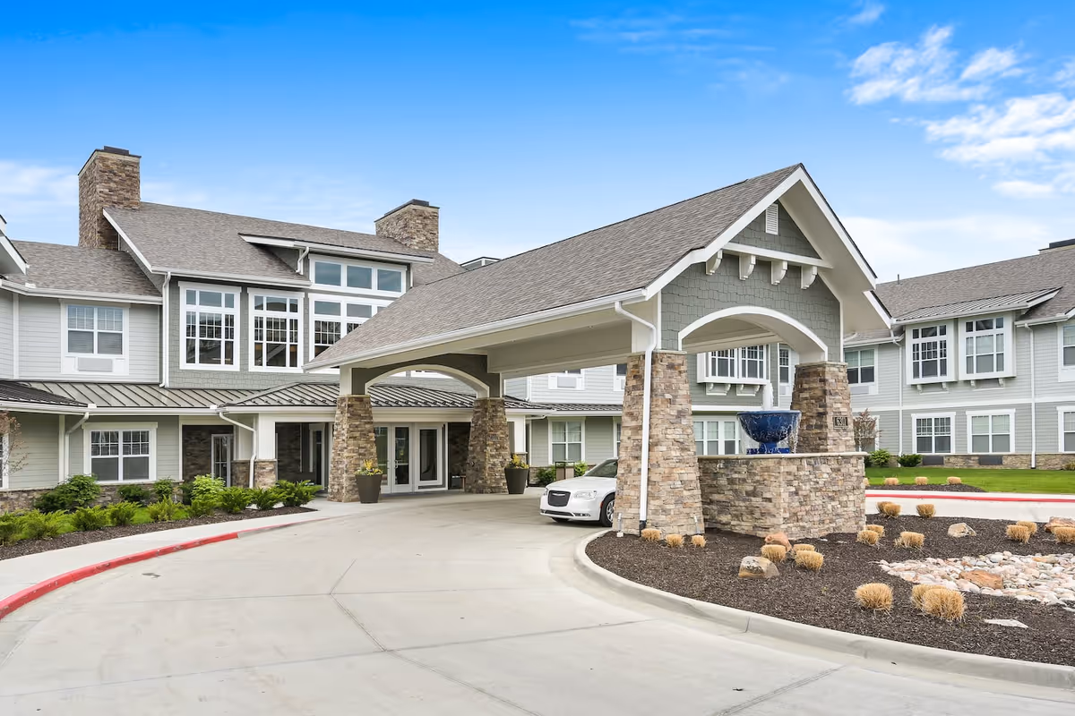 Front exterior view of American House Burlington Creek facility showing a covered entrance with stone pillars, a driveway, landscaped areas with shrubs and rocks, and a blue decorative fountain under the entrance roof.