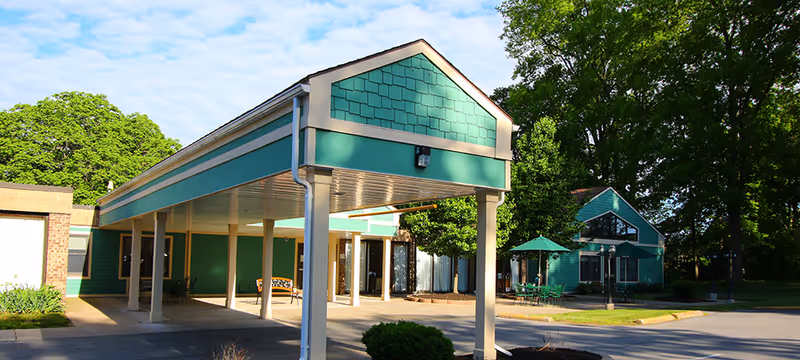 Entrance canopy and front exterior of a teal-painted senior living building with outdoor seating and surrounding trees.