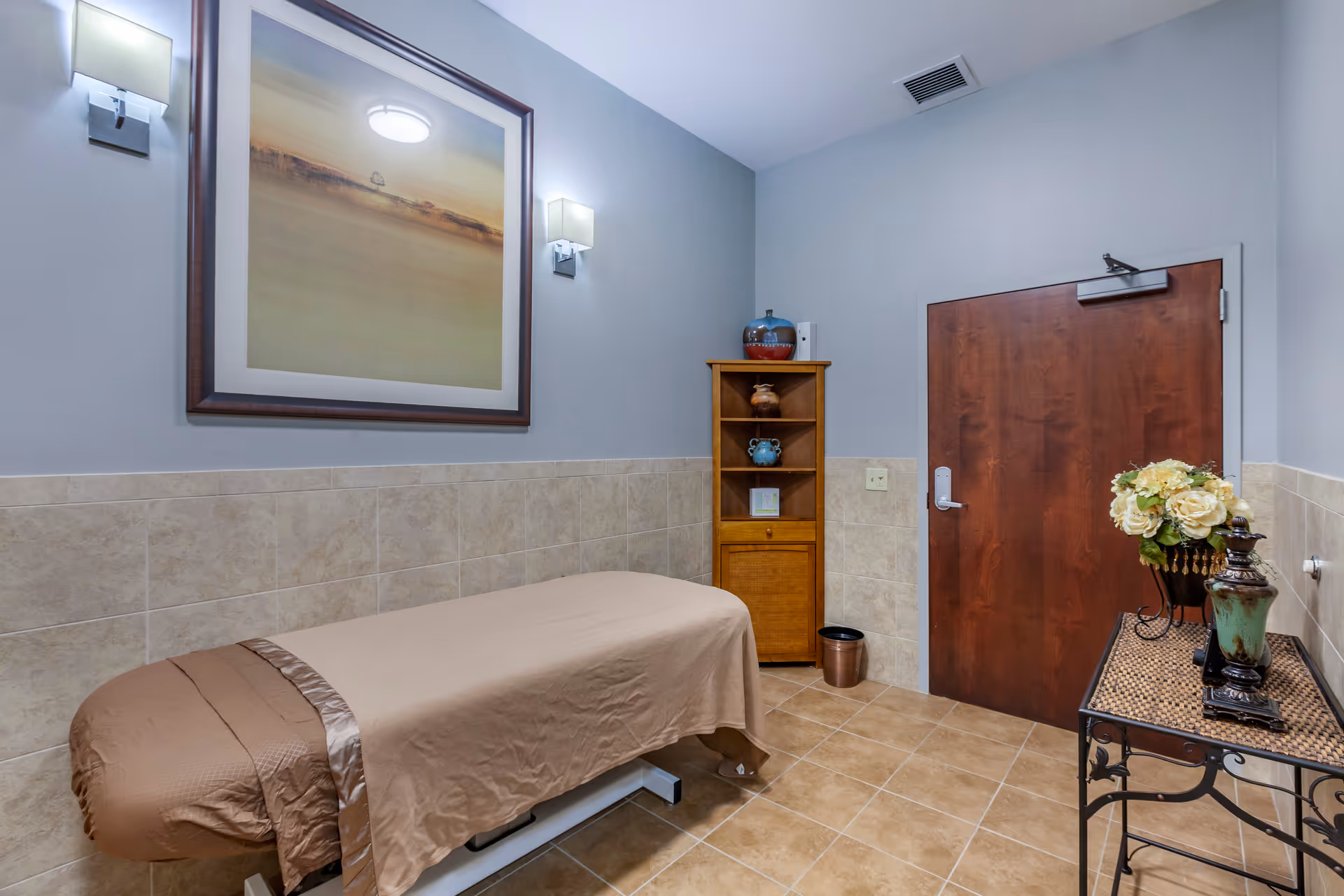 A small treatment room with a massage or examination table covered with a brown sheet, beige tiled floor and half-tiled walls, a wooden cabinet with decorative pottery, a dark wooden door, a metal table with a flower arrangement and decorative vases, and a large framed artwork on the wall illuminated by two wall sconces.