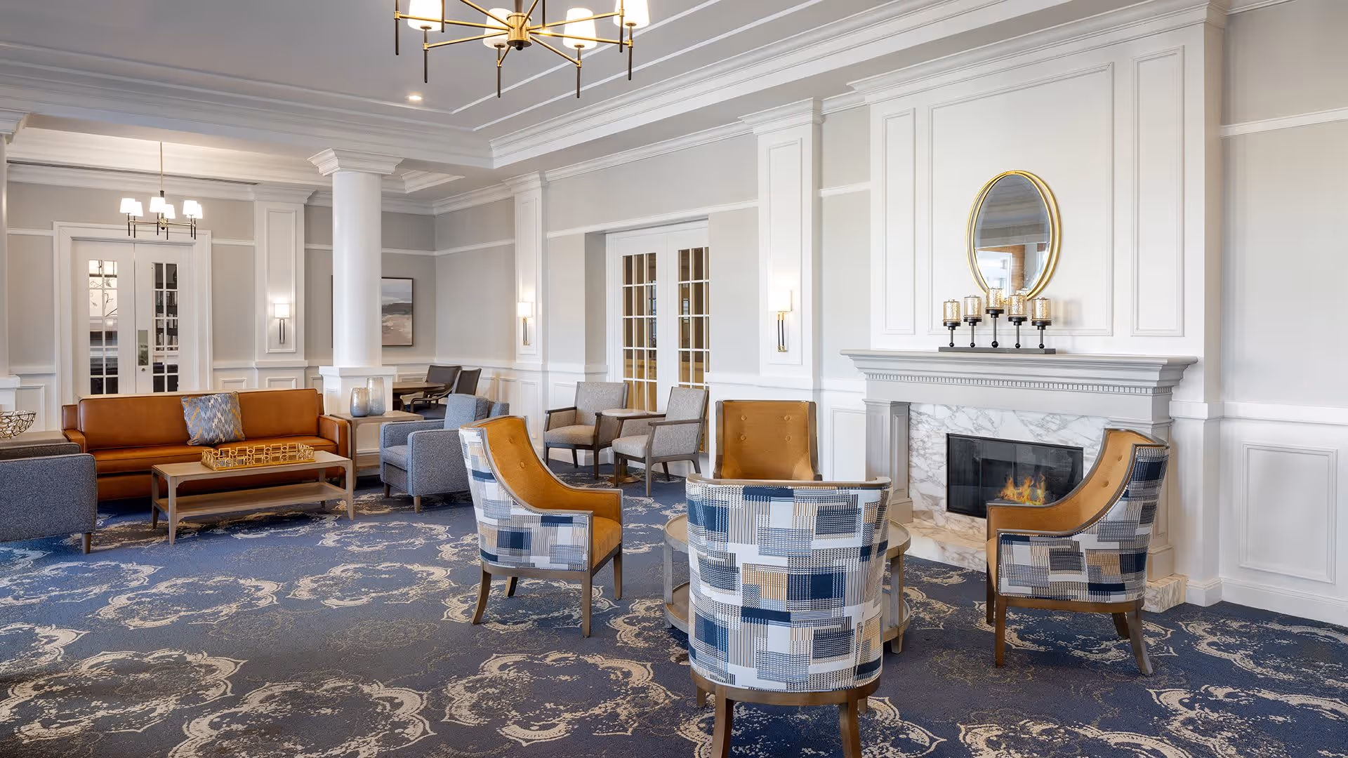 A spacious and elegant living room area in a senior living facility with a blue patterned carpet, a fireplace with a round mirror and candles above it, multiple armchairs with patterned upholstery, a brown leather sofa, and soft lighting from wall sconces and chandeliers.
