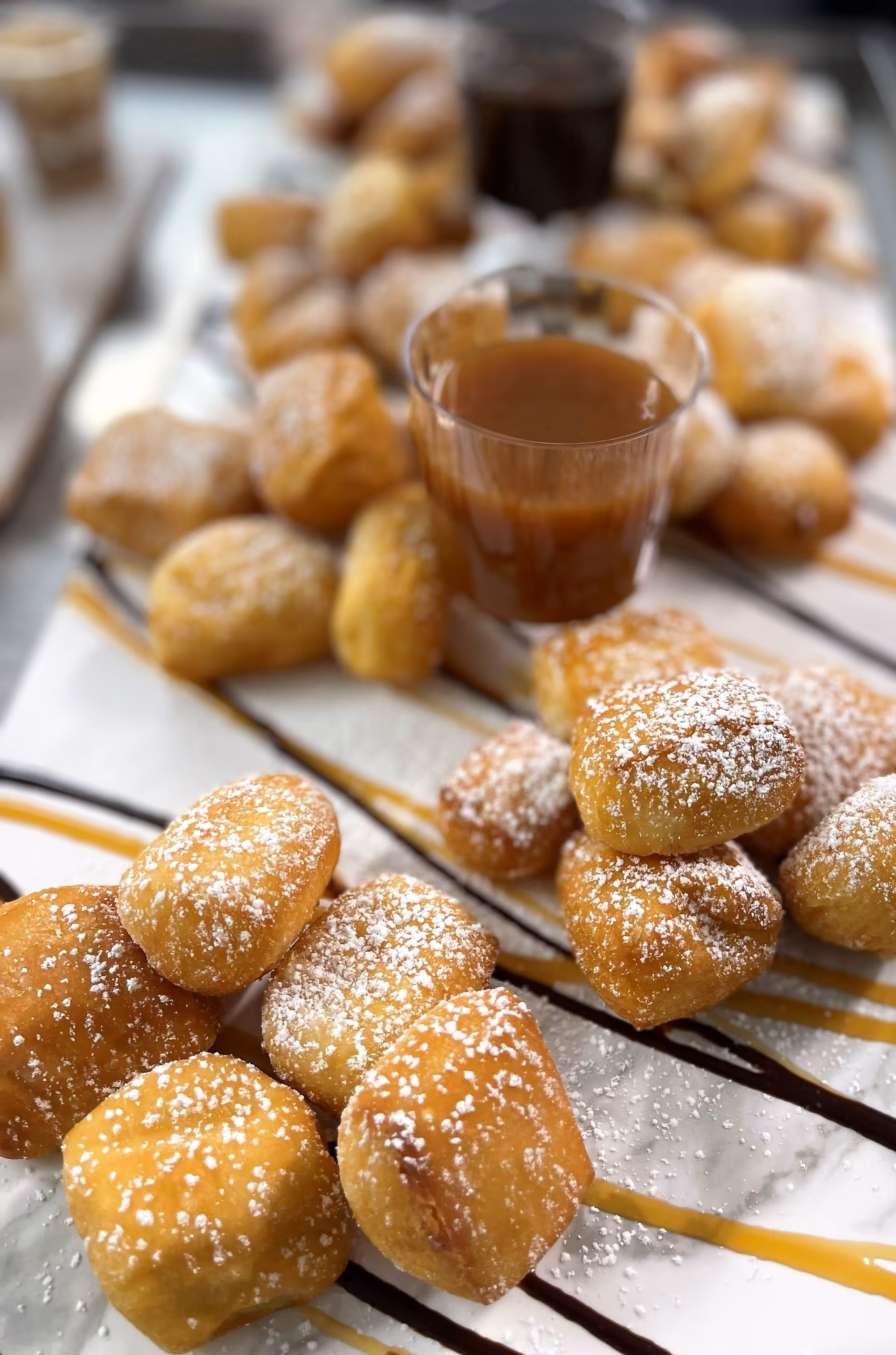 Close-up of powdered sugar-coated fried dough bites arranged on a tray with drizzles of caramel and chocolate sauce, accompanied by small cups of caramel dipping sauce.