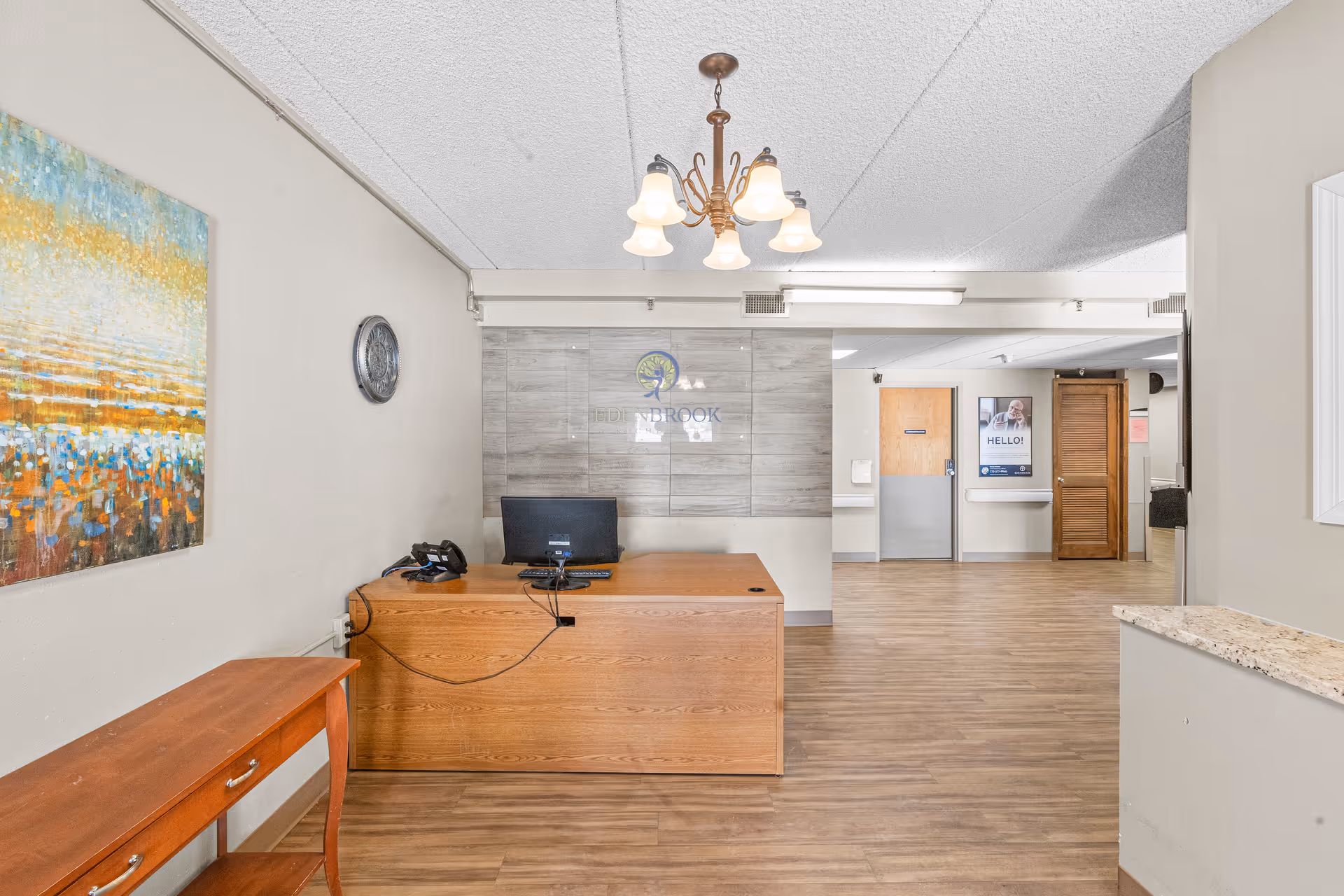 Reception area of Edenbrook Rochester facility with a wooden desk, computer, and phone. There is a colorful abstract painting on the left wall and a clock above it. The floor is wood-patterned, and there is a chandelier hanging from the ceiling. In the background, there are doors and a hallway with a poster on the wall.