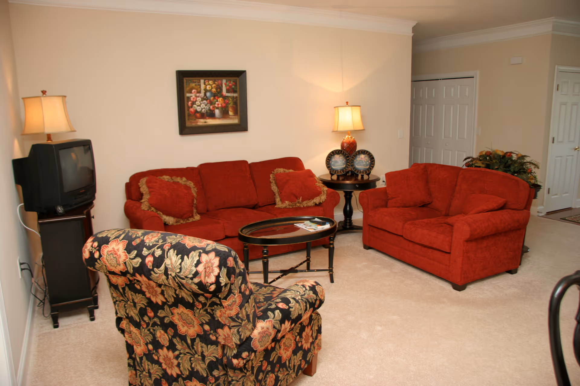 A cozy living room with two red upholstered sofas and a floral patterned armchair arranged around a dark wooden oval coffee table. There are two table lamps on either side of the sofas, one on a small cabinet with an old-style TV and the other on a round side table with decorative plates. A framed floral painting hangs on the wall above the sofa. The room has beige carpet and light-colored walls.