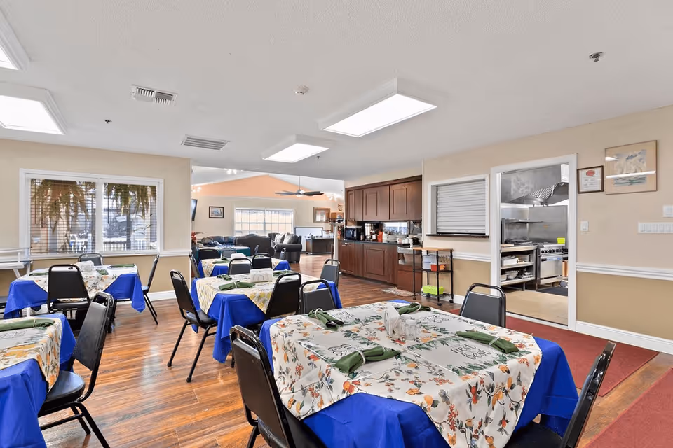 Dining area in a senior living facility with several tables covered in blue and floral tablecloths, each set with green napkins. The room has wooden flooring, beige walls, and large windows letting in natural light. In the background, there is a kitchen area with wooden cabinets and a stainless steel cooking area visible through an open door. Beyond the dining area, a living room with sofas and a ceiling fan can be seen.