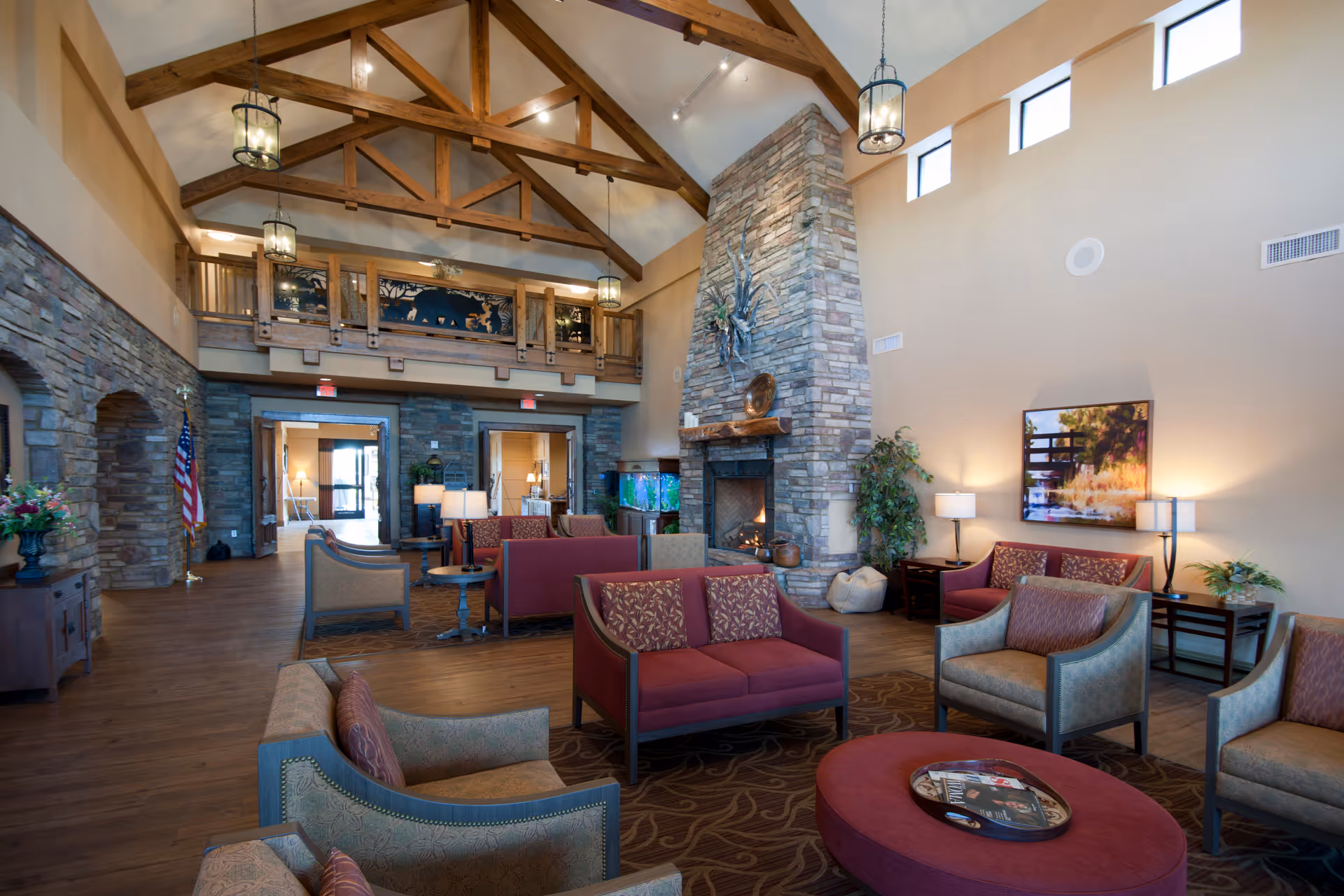 Spacious senior living facility common area with high vaulted ceilings featuring exposed wooden beams, a large stone fireplace, multiple seating arrangements with armchairs and sofas in warm tones, decorative lamps, plants, and artwork on the walls. The room has wooden floors and a balcony overlooking the space.