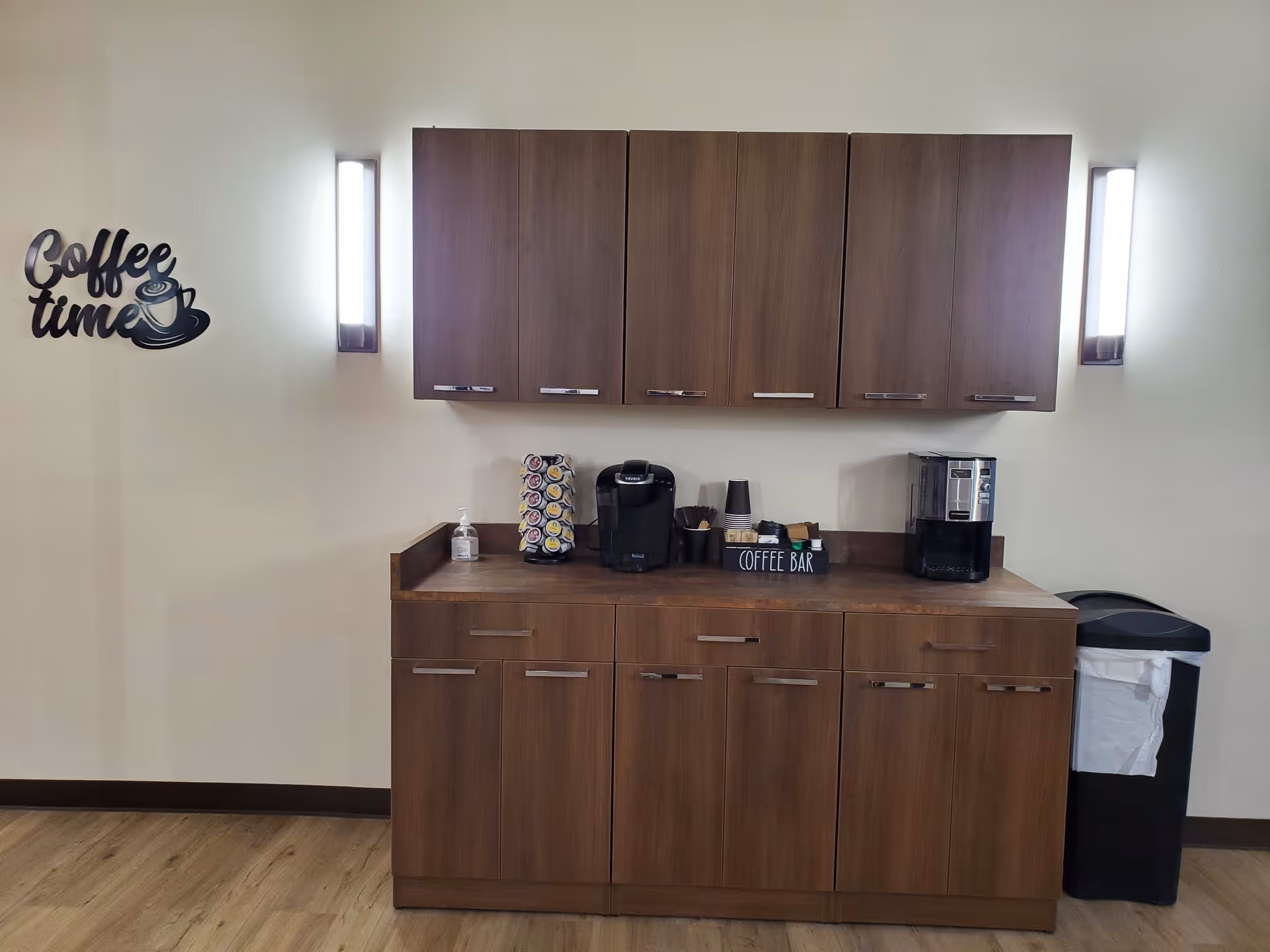 A coffee bar area with wooden cabinets both above and below a countertop. On the countertop, there is a coffee machine, a pod holder with coffee pods, a hand sanitizer bottle, disposable cups, and a small sign that says 'COFFEE BAR'. Two vertical wall lights flank the cabinets. On the left wall, there is a decorative sign that reads 'Coffee time' with a coffee cup graphic. A trash can with a white liner is placed on the right side of the counter.