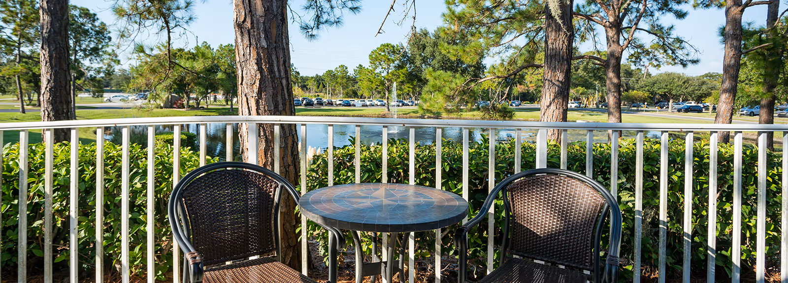 Outdoor patio area with two wicker chairs and a round table, overlooking a pond surrounded by trees and greenery with a parking lot visible in the background.