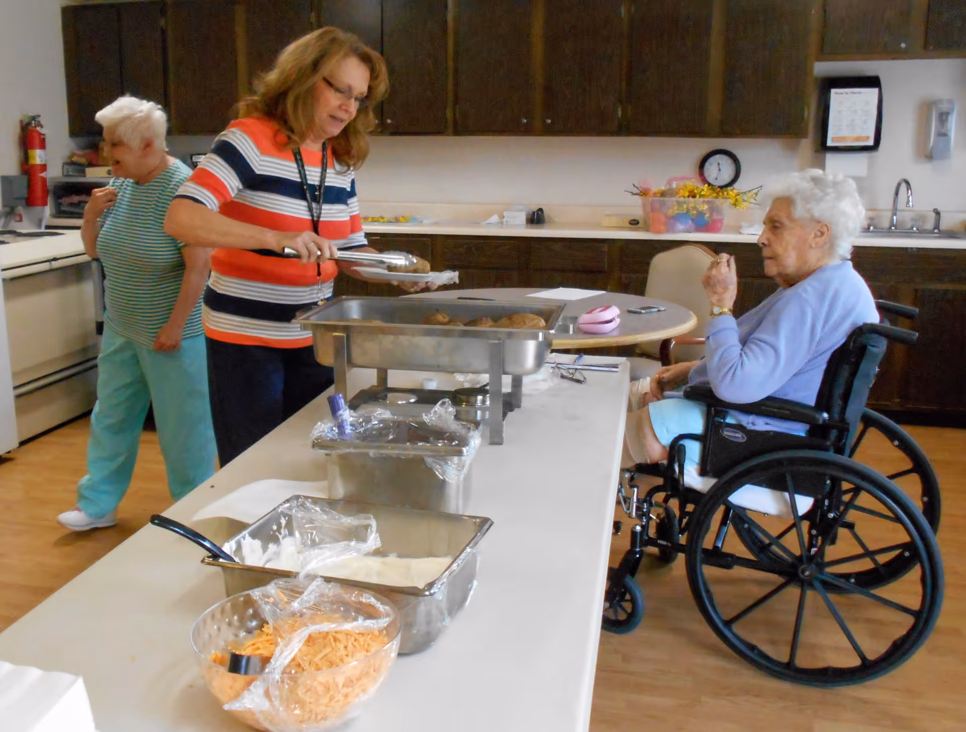 Three elderly women in a kitchen area at Yorktown Manor. One woman in a striped shirt is serving food from a buffet tray, another woman in a wheelchair is sitting at a table eating, and a third woman is standing near the stove in the background. The kitchen has wooden cabinets, a sink, and various food containers on the counter and table.