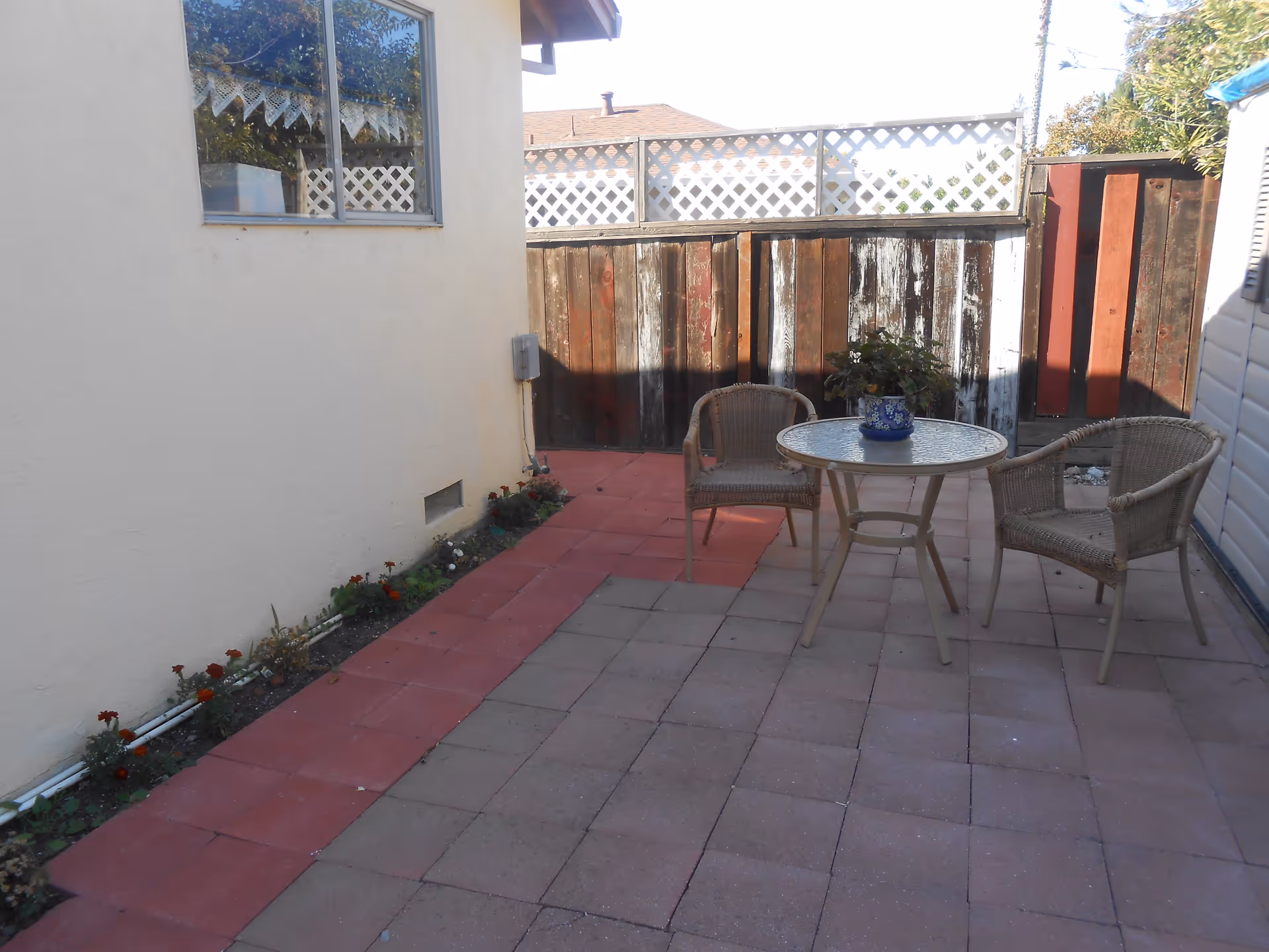 Small enclosed patio with a round glass-top table, two wicker chairs, a potted plant, and a wooden fence.