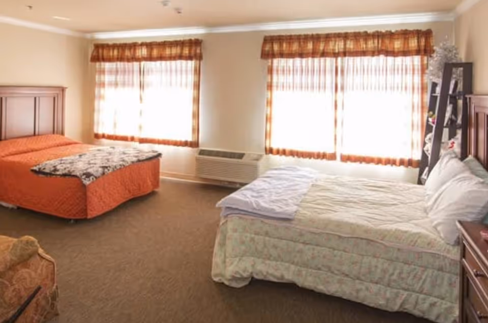A well-lit bedroom with two beds, one with an orange bedspread and the other with a light floral bedspread. The room has two large windows with sheer curtains and valances, a wooden headboard for each bed, a small wooden dresser, and a patterned armchair in the corner.