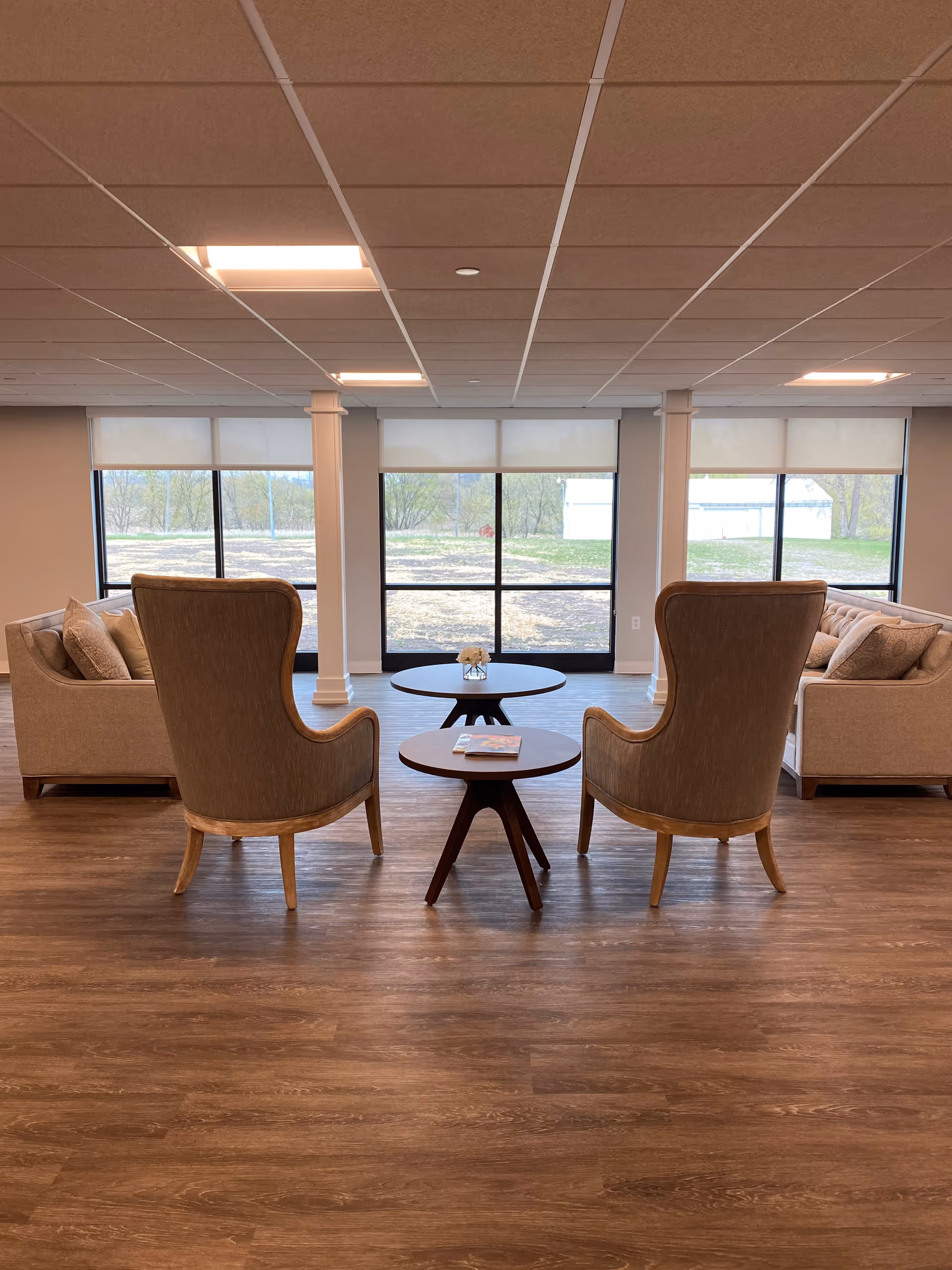 A spacious and well-lit lounge area with two high-backed armchairs facing two beige sofas. Between the chairs are two round wooden tables, one with a small flower arrangement and the other with a book. Large windows in the background let in natural light and offer a view of the outdoors.