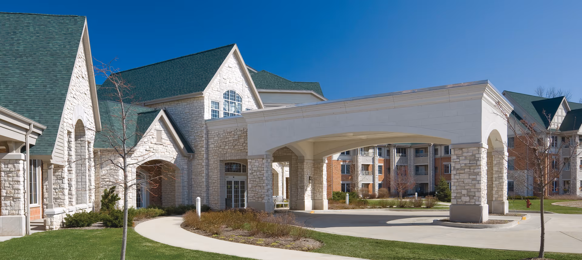 Front exterior of Newcastle Place showing a stone-faced building with green roofs and a covered porte-cochere over the driveway.