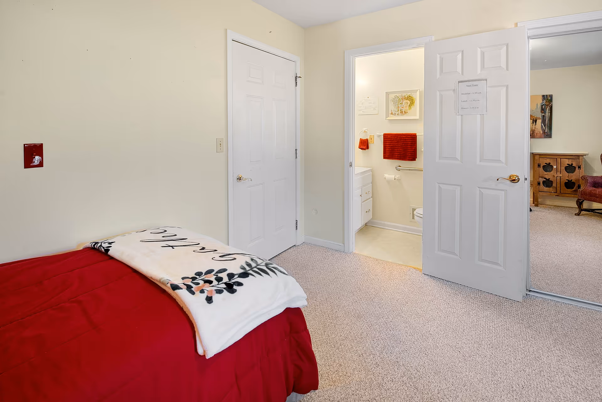 A bedroom with a red bedspread and a white blanket with floral design on the bed. The room has beige walls and carpeted floor. There is a closed white door on the left and an open door on the right leading to a bathroom with a white vanity, red towels, and a toilet. Beyond the bathroom door, part of a living room with a wooden cabinet and a red armchair is visible.