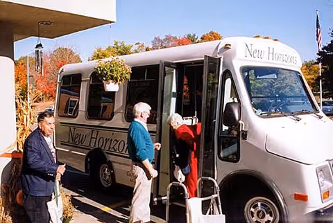 A small white shuttle bus with the name 'New Horizons' on the side and front is parked outside a building. Three elderly individuals are near the bus; one is entering the bus with the assistance of a walker, while the other two stand nearby. The scene is set during autumn with colorful fall foliage in the background.