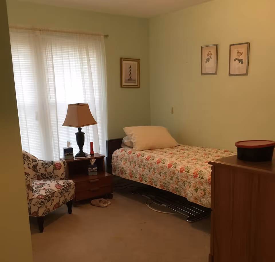 A small bedroom with a single bed covered in a floral quilt, a pillow, a wooden nightstand with a lamp, tissues, and a small red flashlight. There is a floral patterned armchair next to the nightstand, a pair of slippers on the carpeted floor, and three framed pictures on the light green walls. A wooden dresser with a black and red container on top is visible in the foreground.