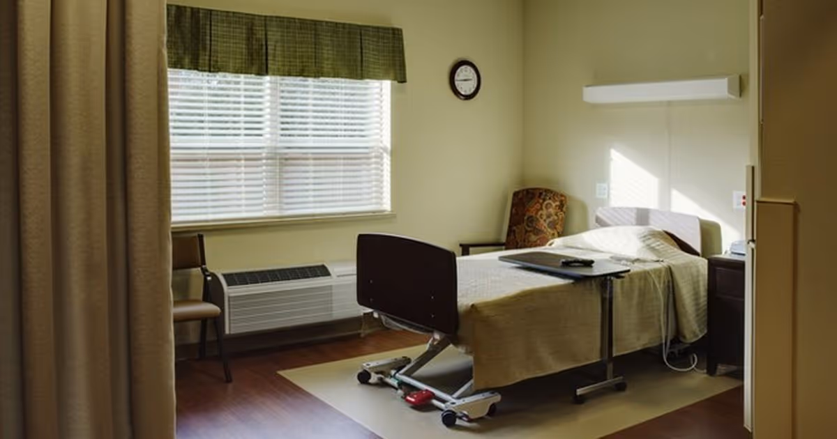 A tidy single-occupancy senior care bedroom with a hospital-style bed, bedside table, armchair, and a window with blinds.