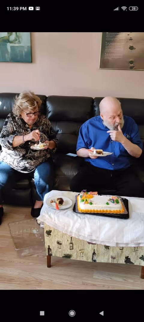 Two elderly individuals sitting on a black leather couch eating cake. A decorated rectangular cake is placed on a small table covered with a white cloth in front of them. The room has light-colored walls with framed artwork hanging.