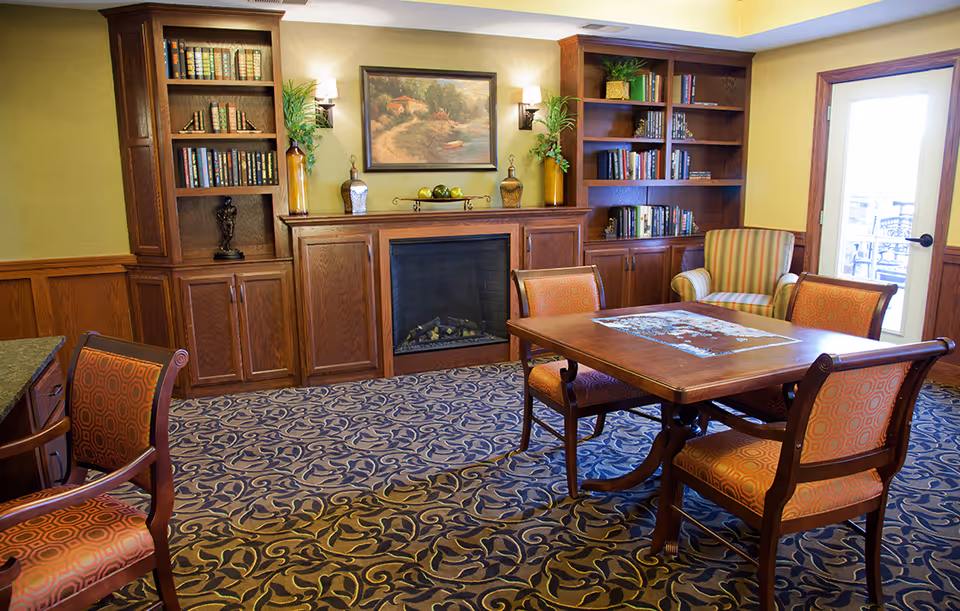 Cozy common room with a table and chairs, a fireplace flanked by built-in bookshelves, and an armchair by a glass door.