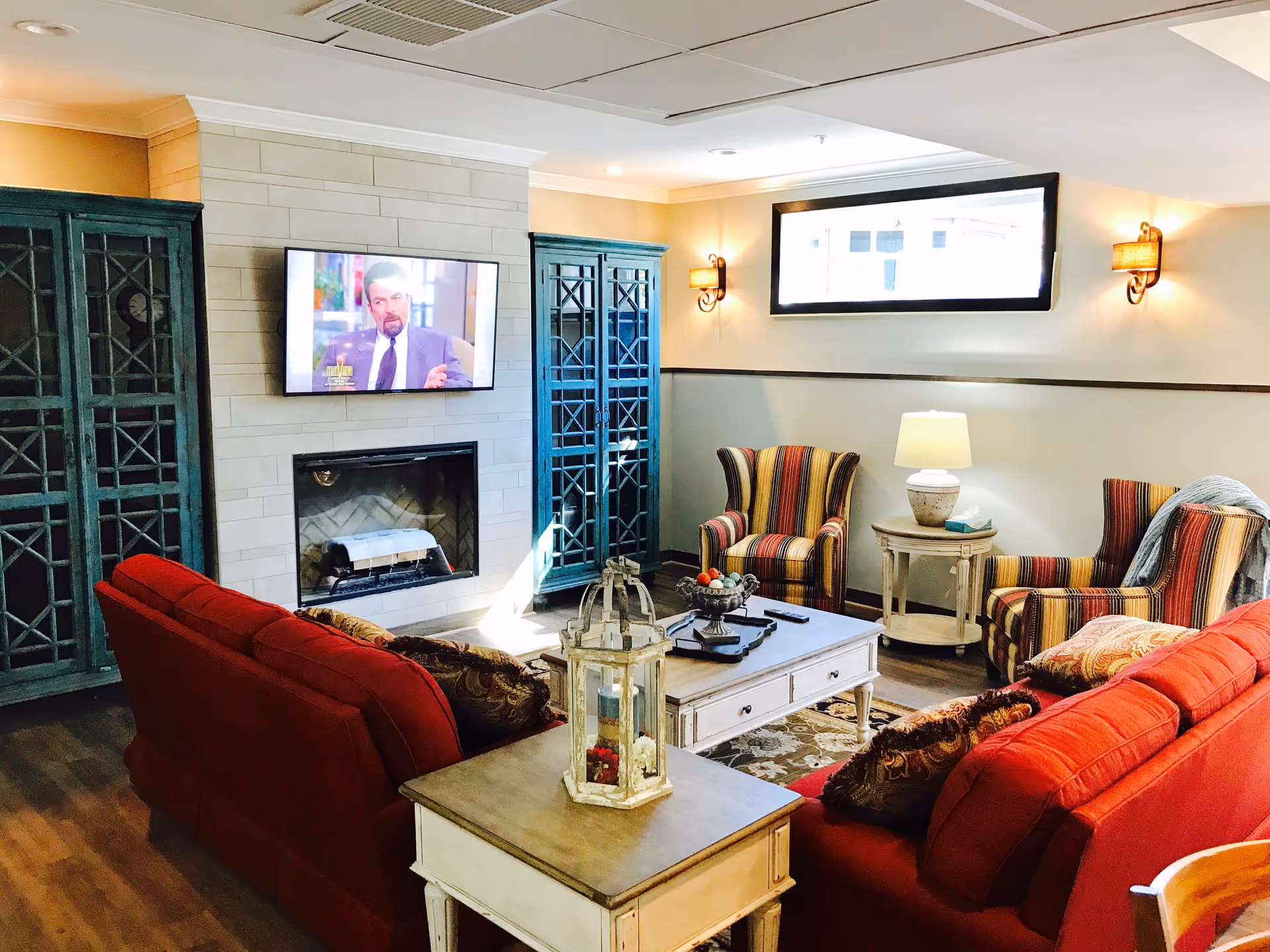 A cozy living room area with two red sofas, two striped armchairs, a white coffee table, and a side table with a lamp. A flat-screen TV is mounted above a fireplace with a light-colored tile surround. Two tall blue cabinets flank the fireplace. The room has wooden flooring and a small window near the ceiling letting in natural light.