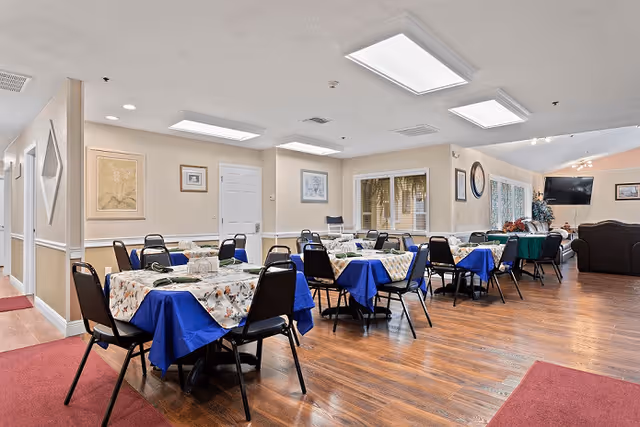 A dining room in a senior living facility with multiple tables covered with floral and blue tablecloths, surrounded by black chairs. The room has wooden flooring, beige walls with framed artwork, and large windows with blinds. There is a television mounted on the far wall and a comfortable seating area with sofas in the background.
