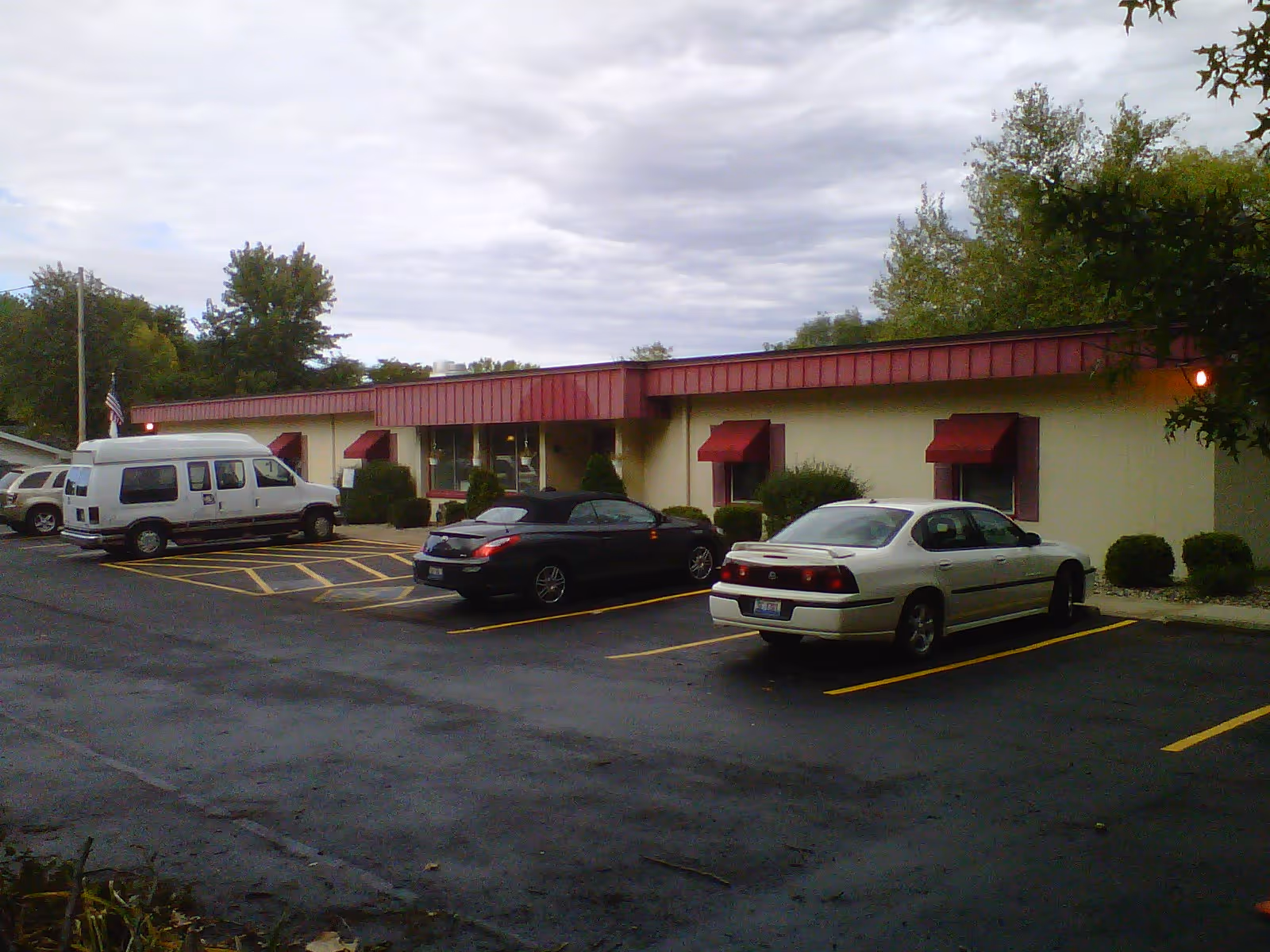 Single-story building with red awnings and several parked cars in a front parking lot under a cloudy sky.